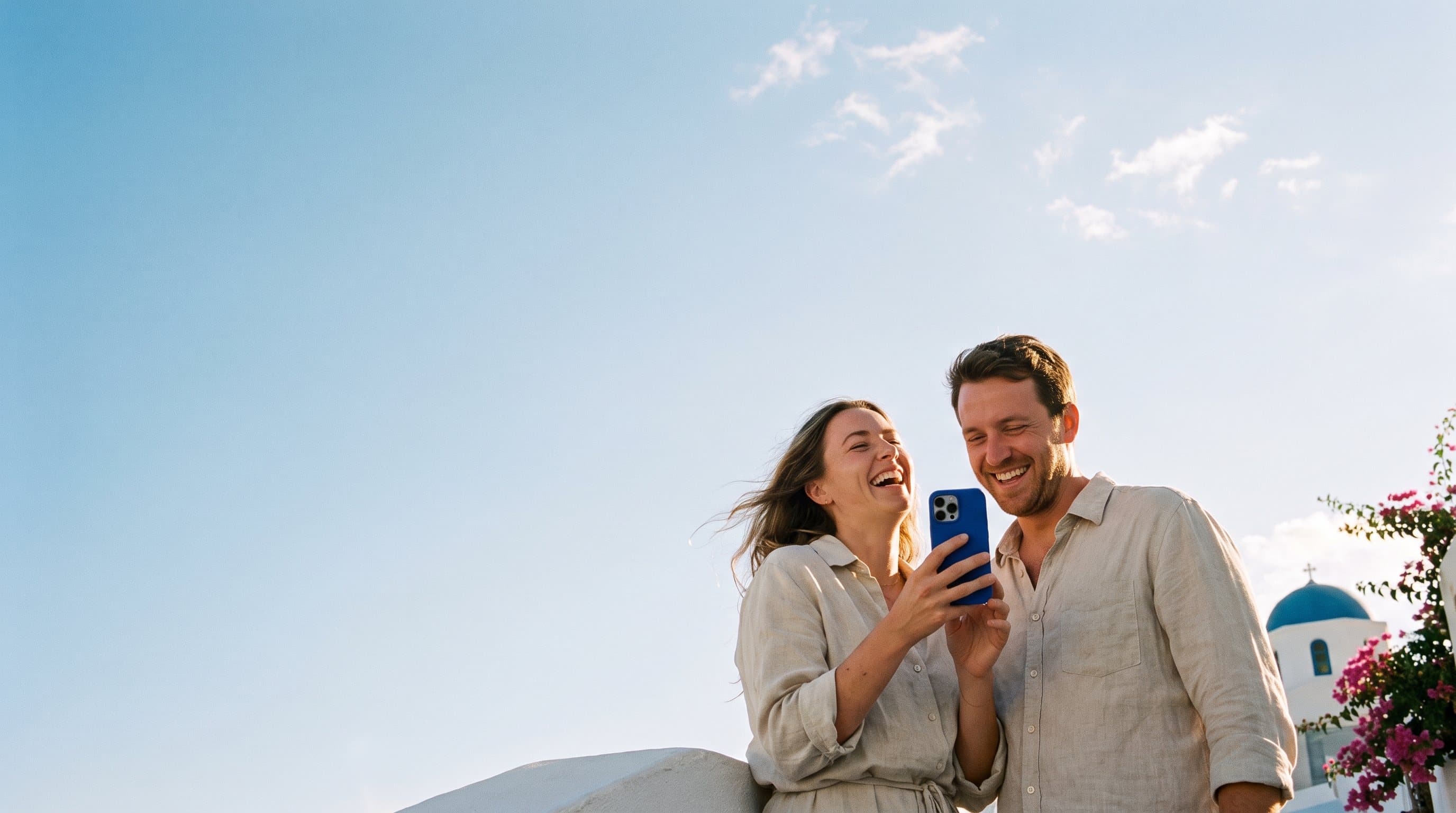 Un couple rit devant un téléphone sur une terrasse blanchie à la chaux, en Grèce, une chapelle au dôme bleu derrière lui